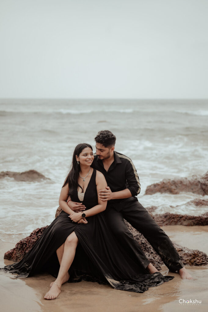 A cute couple wearing black dresses sitting on a big rock near beach picture captured by the best pre-wedding photographer in Delhi