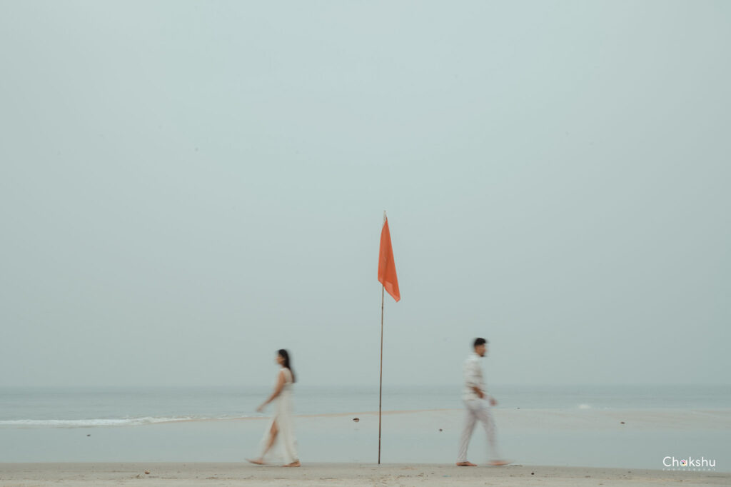 A couple strolls along the beach, an orange flag waving nearby, capturing a moment perfect for a pre-wedding photographer in Delhi.